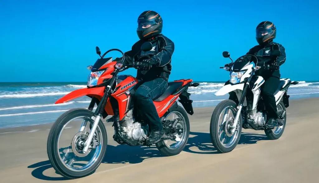 dois pilotos em moviemnto em suas motocicletas honda nxr 160 bros uma vermelha e outra branca a beira mar - Moto Honda Motopel